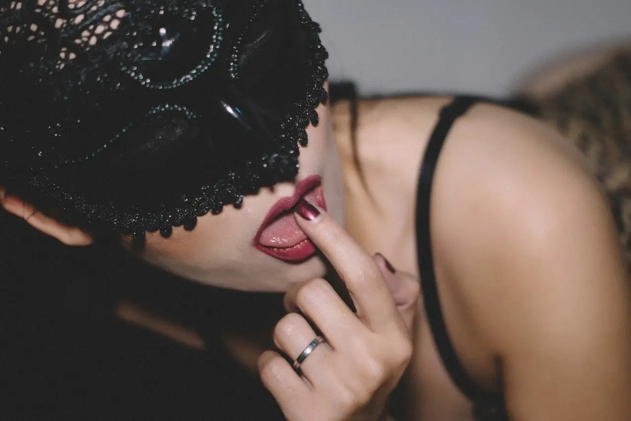 Close-up of a sensual woman wearing a black lace mask and touching her lips suggestively, symbolising fantasy, desire, and the art of dirty talk.