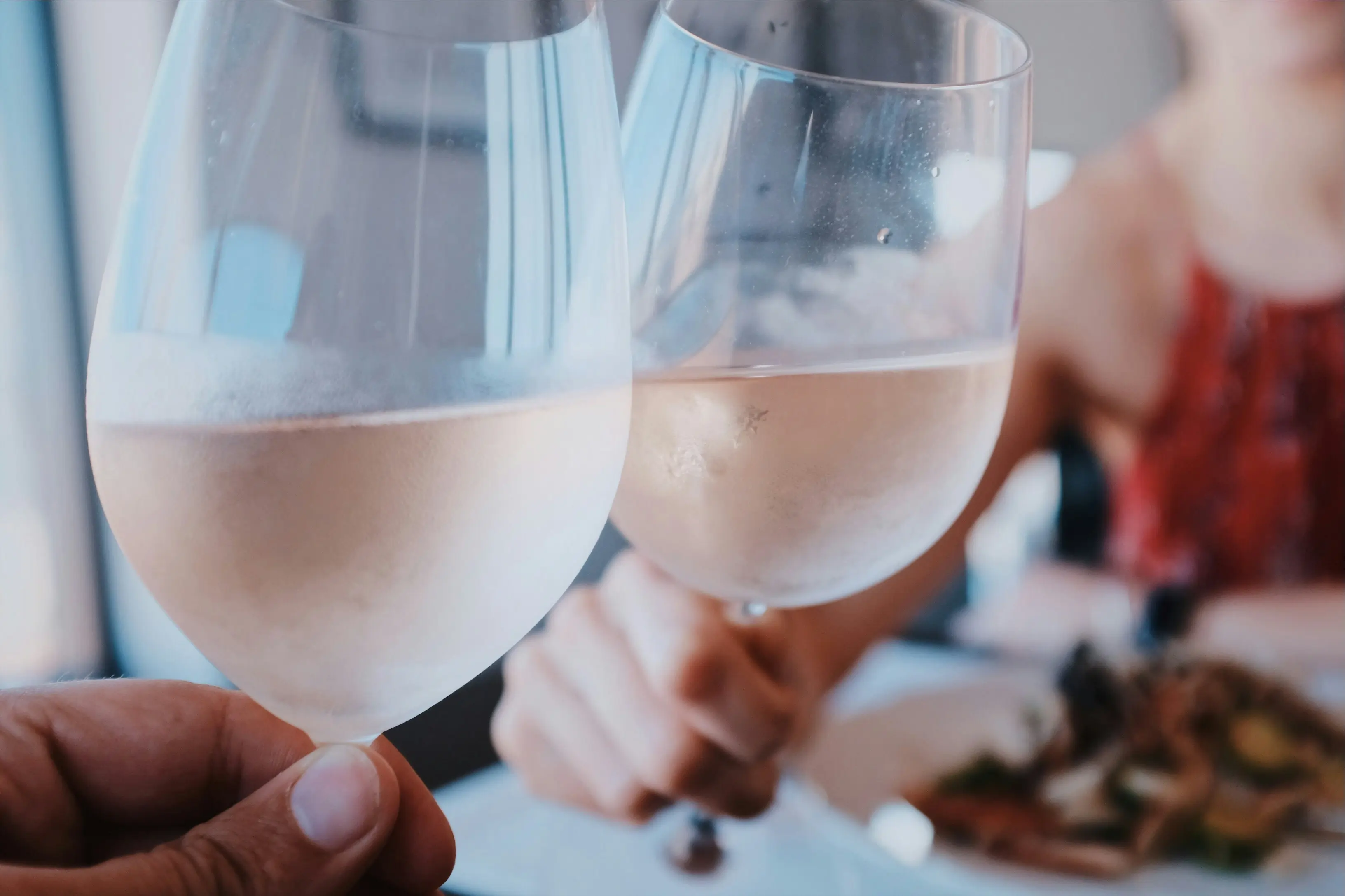 Close-up of two people clinking glasses of chilled white wine during a dinner date, with food in the background and soft natural lighting.