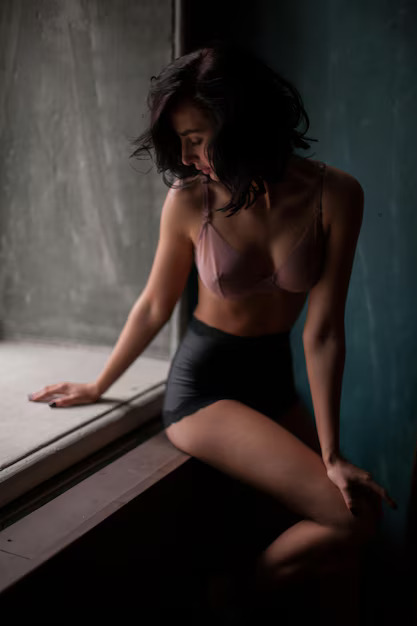 Brunette woman in lingerie sitting by a window in soft light, looking down with a sensual and introspective expression — Angels Escorts atmosphere.