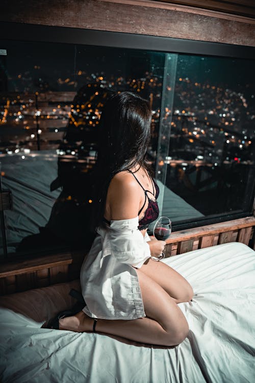 Femme sensuelle en lingerie et chemise blanche assise sur un lit, tenant un verre de vin et regardant la ville illuminée à travers la fenêtre d’un hôtel.